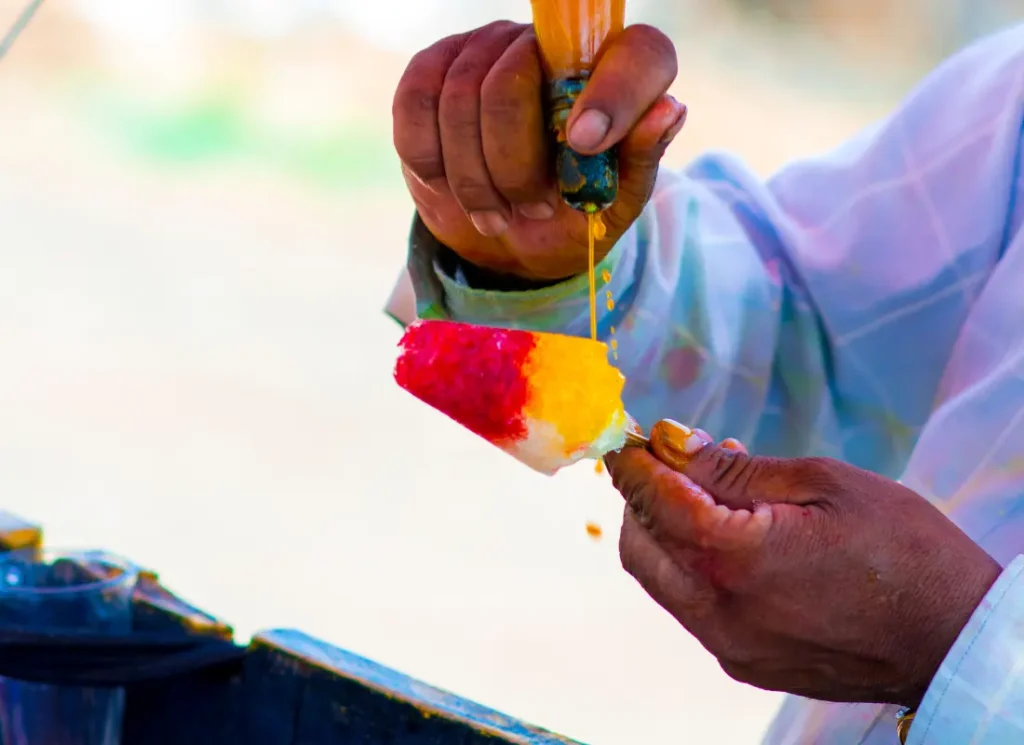 a man putting red and orange coloured syrups onto an Indian gola